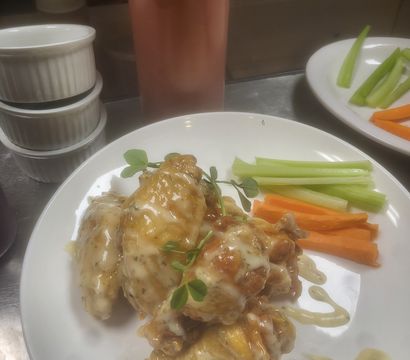 Sticky glazed chicken wings with creamy herb drizzle and microgreens, served with celery and carrot sticks on a white plate in a commercial kitchen.