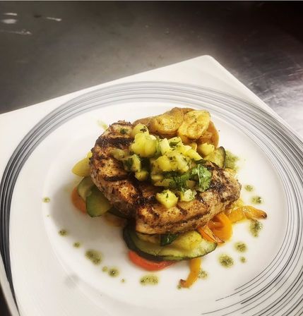 Grilled charred chicken breast topped with bright pineapple-cilantro salsa, served on sautéed zucchini and bell peppers with crispy baby potatoes and herb-oil dots on a white plate.