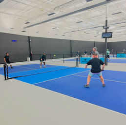 Indoor pickleball doubles match on bright blue courts, four players positioned at the net with a ball midair in a modern indoor sports facility.