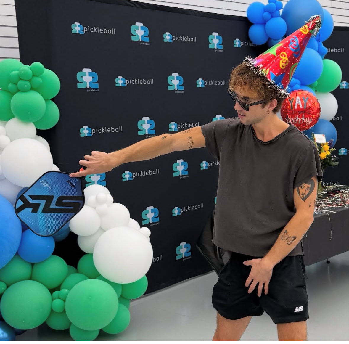Person in sunglasses and a colorful party hat holding a blue pickleball paddle at an indoor pickleball-themed birthday celebration with green, blue and white balloon arch and backdrop.