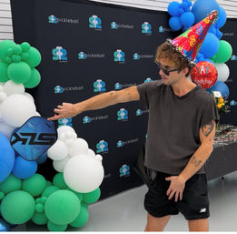 Person in sunglasses and a colorful party hat holding a blue pickleball paddle at an indoor pickleball-themed birthday celebration with green, blue and white balloon arch and backdrop.