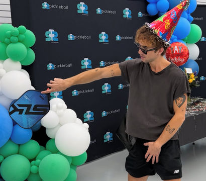 Person in sunglasses and a colorful party hat holding a blue pickleball paddle at an indoor pickleball-themed birthday celebration with green, blue and white balloon arch and backdrop.