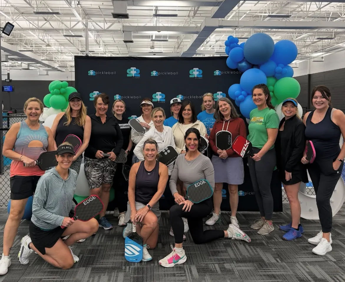 Smiling group of women pickleball players posing with paddles in front of a branded backdrop and blue-green balloon arch inside an indoor pickleball facility.