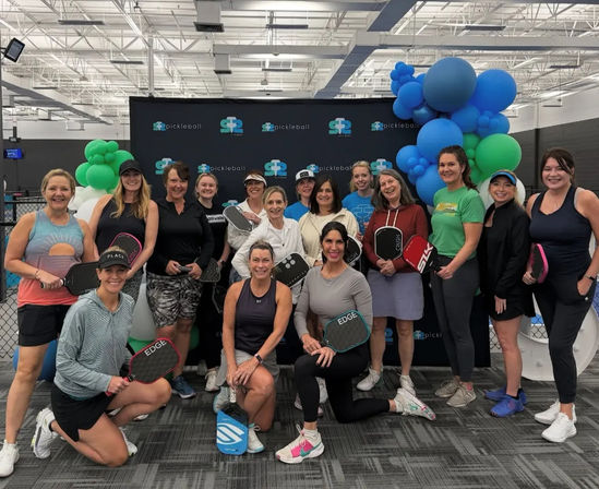 Smiling group of women pickleball players posing with paddles in front of a branded backdrop and blue-green balloon arch inside an indoor pickleball facility.
