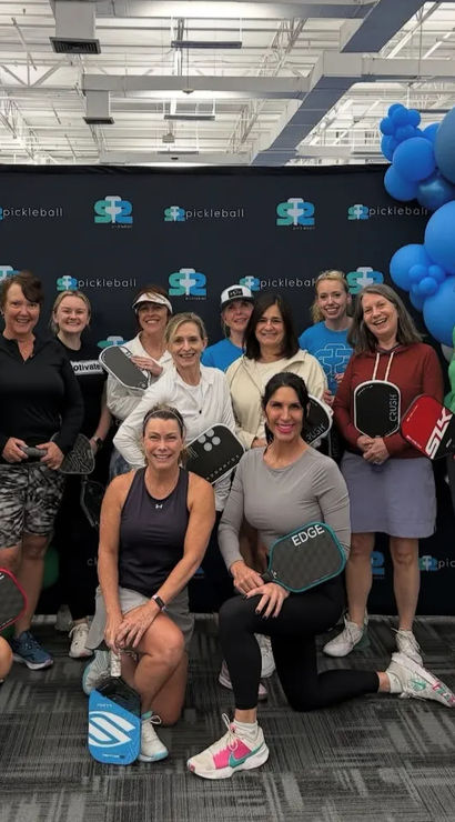 Smiling group of women pickleball players posing with paddles in front of a branded backdrop and blue-green balloon arch inside an indoor pickleball facility.
