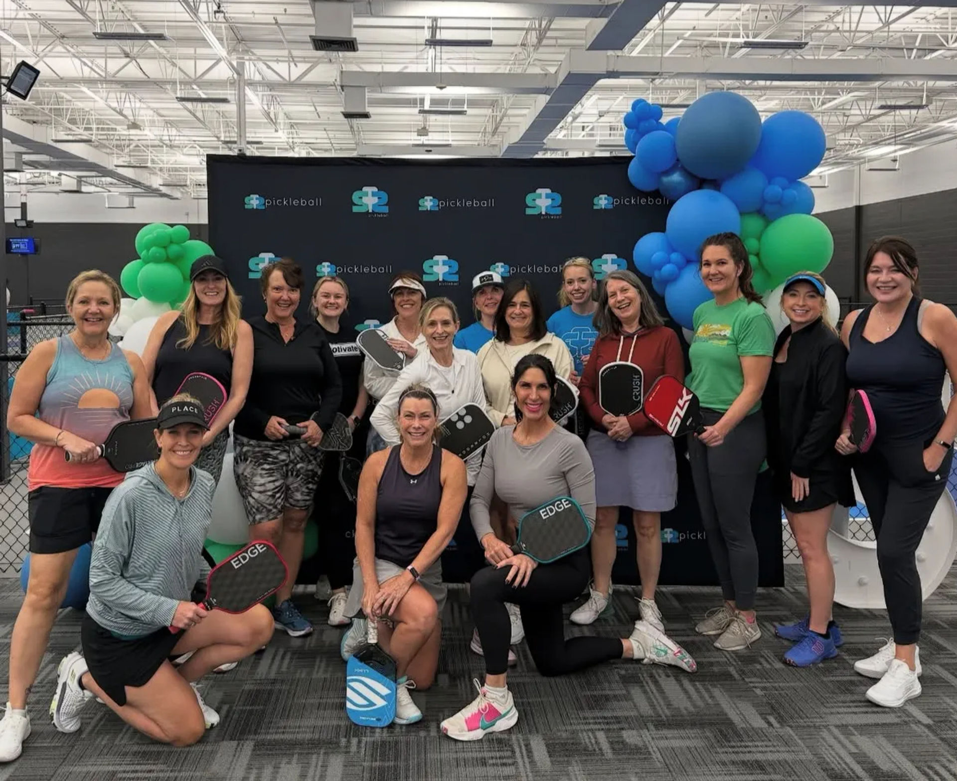 Smiling group of women pickleball players posing with paddles in front of a branded backdrop and blue-green balloon arch inside an indoor pickleball facility.