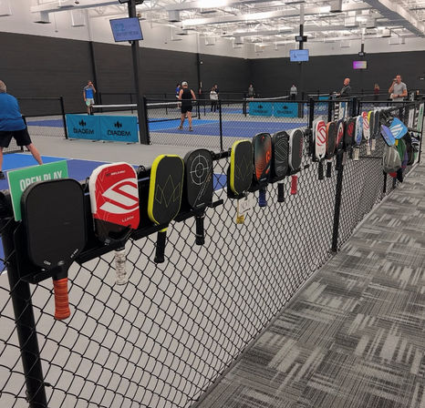 Colorful pickleball paddles hung on a chain-link fence overlooking multiple indoor blue pickleball courts with players in action under bright arena lighting