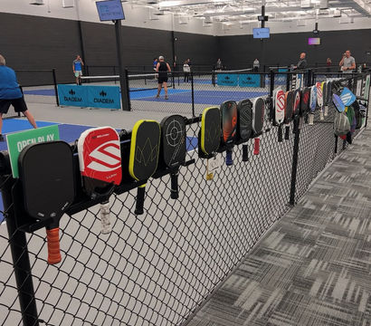 Colorful pickleball paddles hung on a chain-link fence overlooking multiple indoor blue pickleball courts with players in action under bright arena lighting