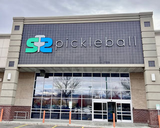 Modern pickleball facility storefront with teal-and-blue paddle logo and large "pickleball" sign on a tiled facade above mirrored glass doors reflecting trees and parked cars.