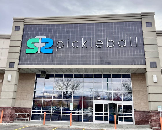 Modern pickleball facility storefront with teal-and-blue paddle logo and large "pickleball" sign on a tiled facade above mirrored glass doors reflecting trees and parked cars.
