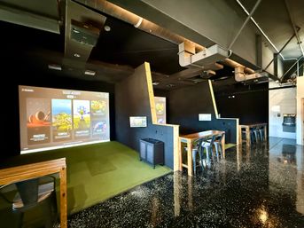 Indoor golf simulator lounge with a large projected game menu over green turf hitting bays, wooden high-top tables and metal stools, exposed industrial ductwork and glossy speckled floor.