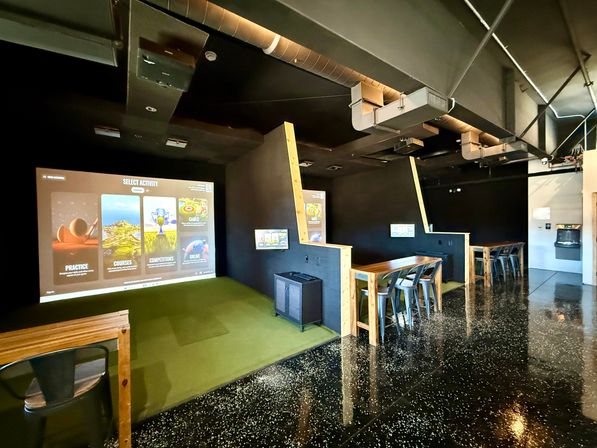 Indoor golf simulator lounge with a large projected game menu over green turf hitting bays, wooden high-top tables and metal stools, exposed industrial ductwork and glossy speckled floor.