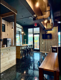 Modern cafe interior with wooden bar and high stools, pendant lights, striped wood accent wall, glossy speckled floor, and large windows showing an evergreen forest view.