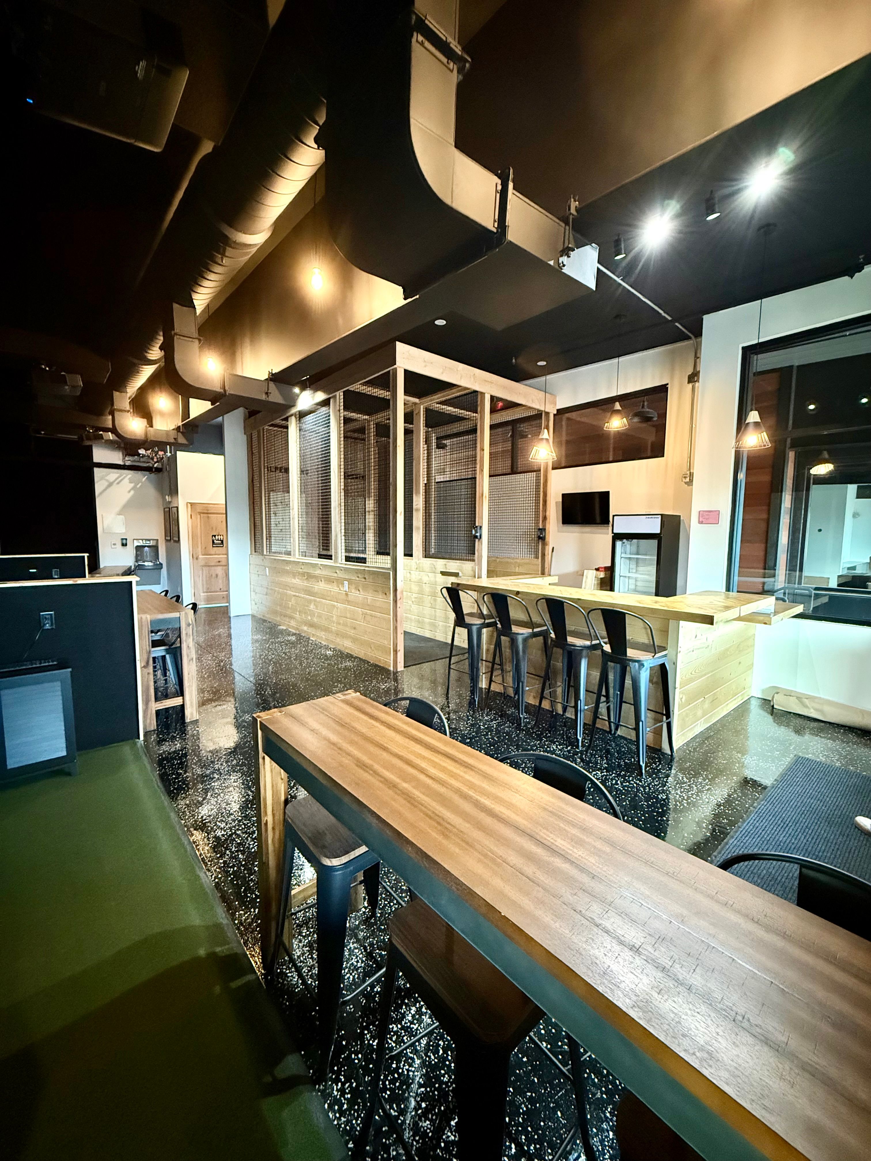 Industrial-style communal lounge with high black ceiling and exposed ductwork, long wooden tables and bar with metal stools, wood-and-wire mesh partition, pendant lights and speckled polished black floor.