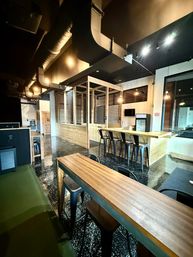 Industrial-style communal lounge with high black ceiling and exposed ductwork, long wooden tables and bar with metal stools, wood-and-wire mesh partition, pendant lights and speckled polished black floor.