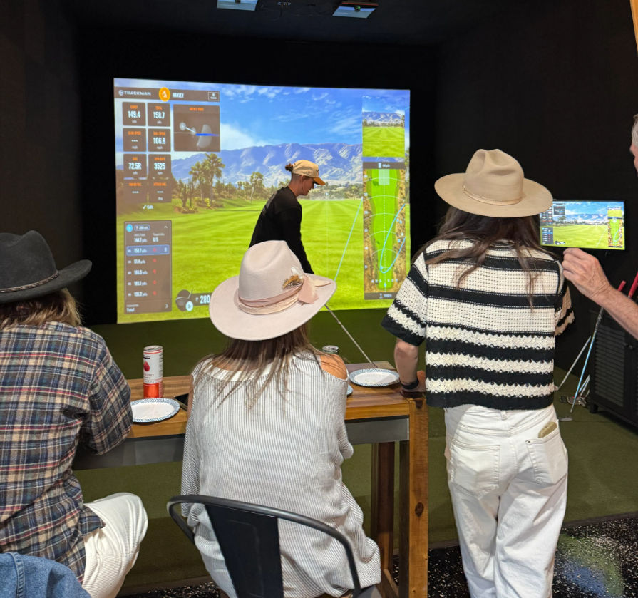 Friends wearing hats play an indoor golf simulator — a player swings toward a large projected virtual fairway with shot-tracking stats while others watch from a wooden counter with drinks.