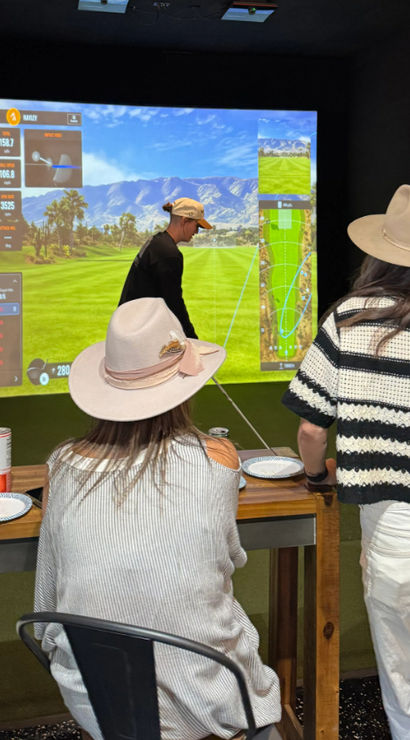 Friends wearing hats play an indoor golf simulator — a player swings toward a large projected virtual fairway with shot-tracking stats while others watch from a wooden counter with drinks.