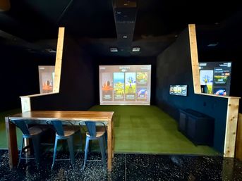 Indoor golf simulator bay with a large projection screen showing game menus, green turf hitting area, wooden high table with three metal bar stools, dark walls and ceiling — virtual golf practice setup.