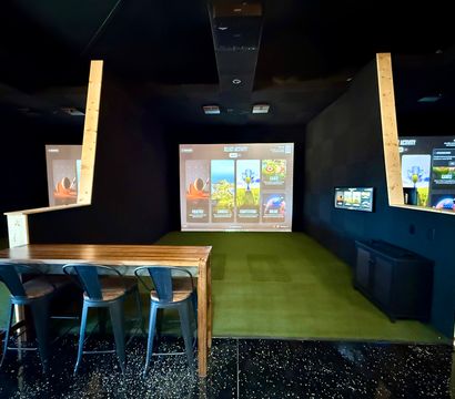Indoor golf simulator bay with a large projection screen showing game menus, green turf hitting area, wooden high table with three metal bar stools, dark walls and ceiling — virtual golf practice setup.
