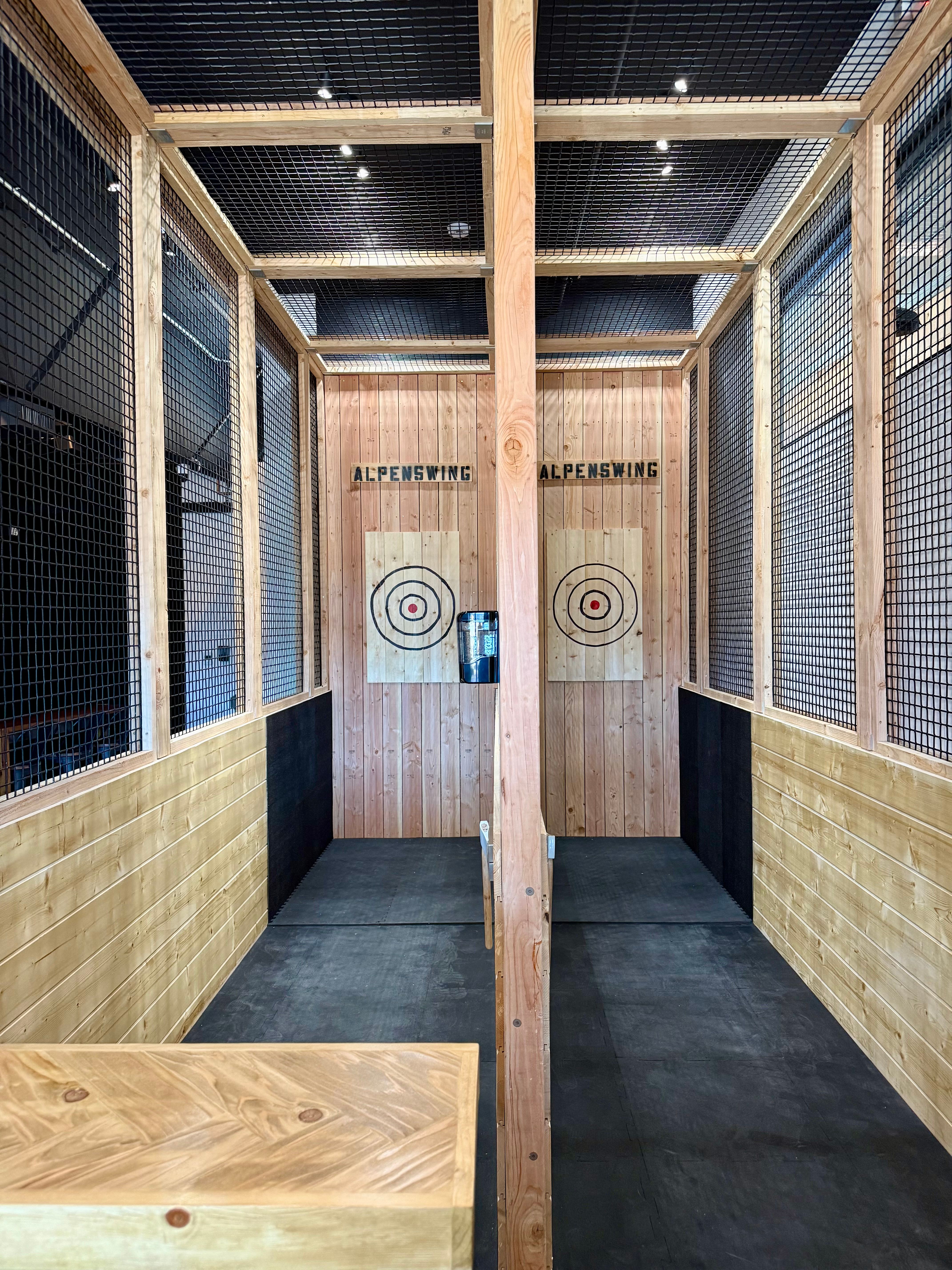 Indoor axe-throwing lanes with wood-paneled walls, safety netting, rubber flooring and two bullseye target boards ready for throwing.