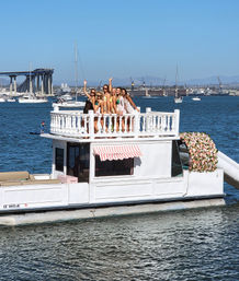 Six friends posing and waving on the upper deck of a white, flower-adorned party boat in a sunny bay with sailboats and a distant bridge