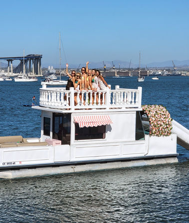 Six friends posing and waving on the upper deck of a white, flower-adorned party boat in a sunny bay with sailboats and a distant bridge