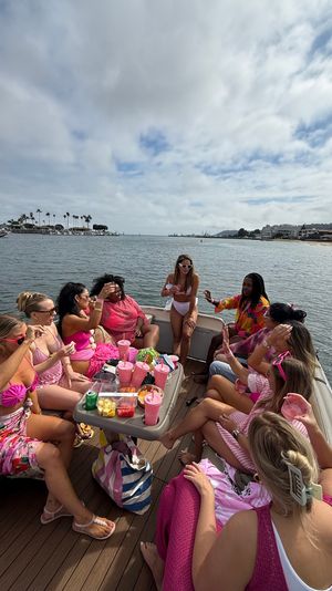 Boat party with a group of women in pink swimsuits sharing drinks and snacks on a palm-lined coastal bay under a cloudy sky.