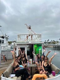 Group of women partying on a docked party boat at a marina under a cloudy sky; one woman in white poses on the upper deck while friends in black swimwear cheer from the lower seating area with palm trees and other boats visible.