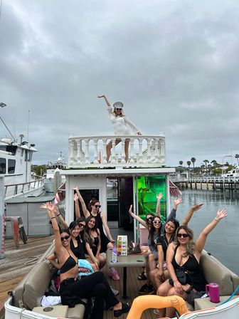 Group of women partying on a docked party boat at a marina under a cloudy sky; one woman in white poses on the upper deck while friends in black swimwear cheer from the lower seating area with palm trees and other boats visible.