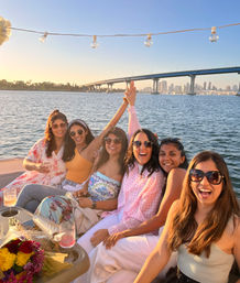 Six friends laughing on a sunset boat cruise in San Diego Bay, string lights overhead, Coronado Bridge and downtown skyline in the background, with drinks and a bouquet in the foreground.
