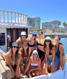 Eight women at a sunny bachelorette boat party on a marina, wearing swimsuits, sun hats and sunglasses with waterfront condos and palm trees in the background