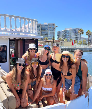 Eight women at a sunny bachelorette boat party on a marina, wearing swimsuits, sun hats and sunglasses with waterfront condos and palm trees in the background