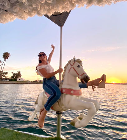 Smiling woman in sunglasses riding a white carousel horse over coastal water at sunset with palm trees and a marina in the background