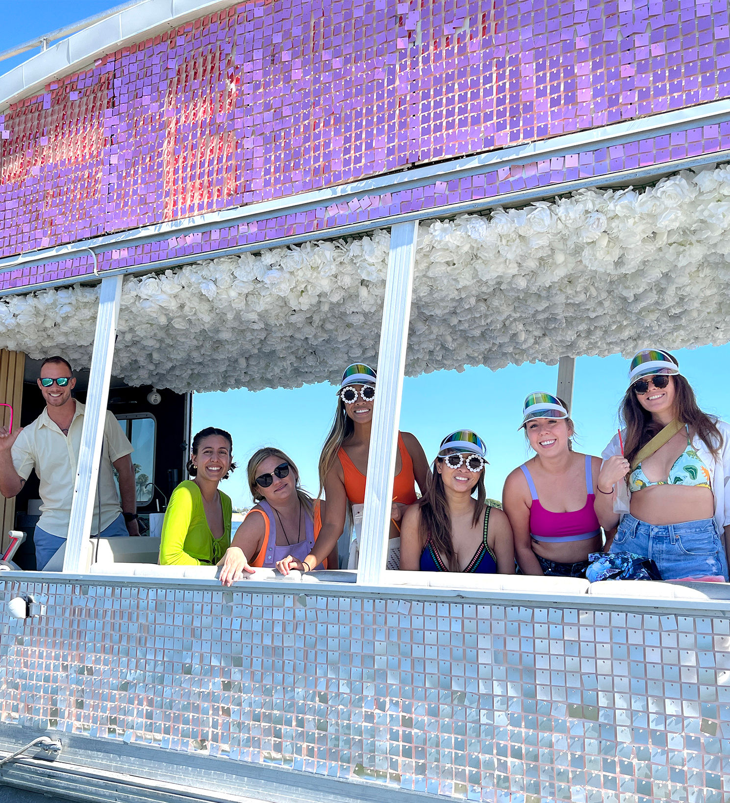 Group of seven adults in colorful swimwear and mirrored visors smiling from a sparkly purple-and-silver decorated party float with a white floral ceiling against a clear blue sky — summer beachside celebration.