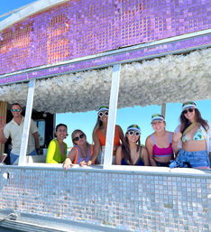 Group of seven adults in colorful swimwear and mirrored visors smiling from a sparkly purple-and-silver decorated party float with a white floral ceiling against a clear blue sky — summer beachside celebration.