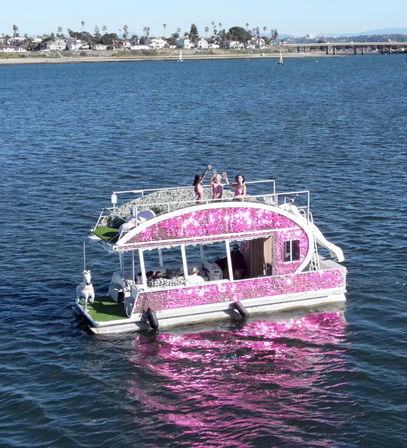 Pink sequined pontoon party boat with people toasting on the upper deck, sparkling reflection on bay waters with coastal homes and a bridge in the background.
