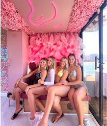 Four women in bikinis posing on a pink-feathered lounge inside a boat, neon pink ceiling light art above and marina skyline visible through the window — playful summer girls’ trip vibe.