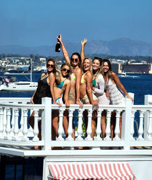 Six women in colorful swimsuits celebrating with a champagne bottle on a white waterfront balcony overlooking a marina with yachts, harbor buildings and distant hills on a sunny summer day.