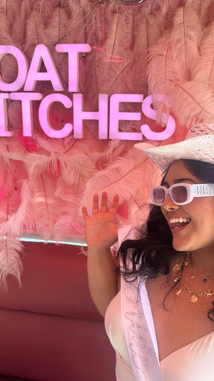 Playful bachelorette photo on a boat: smiling woman in a white cowboy hat and 'BRIDE' sunglasses posing under a pink neon "BOAT BITCHES" sign against a feathery pink backdrop