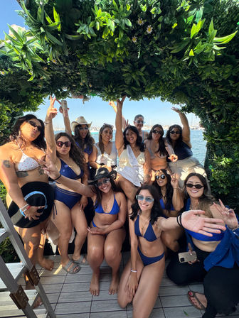 Group of friends in bikinis posing and cheering under a leafy arch on a sunny marina dock with sailboats in the background