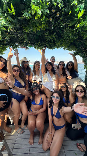 Smiling group of women in bikinis enjoying a sunny boat party under a leafy green arch, cheering with drinks at a marina for a fun bachelorette-style outing.