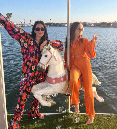Two friends barefoot on a white carousel horse mounted on a boat deck at a sunny marina — one in a bold floral jumpsuit, the other in a bright orange two-piece flashing a peace sign; bubbles, palm trees and waterfront homes in the golden-hour background.