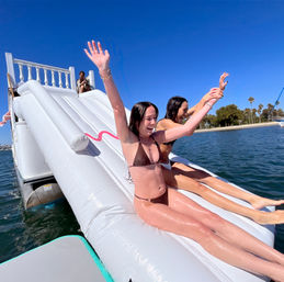 Two women in bikinis with arms raised sliding down an inflatable water slide off a pontoon boat into a sunny coastal bay with palm trees and clear blue sky