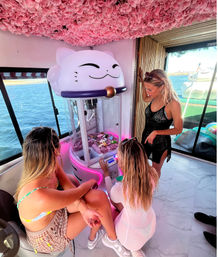 Three women in swimsuits play a giant cat-shaped claw machine with pink neon lights beneath a floral pink ceiling aboard a yacht, ocean and dock visible through panoramic windows.