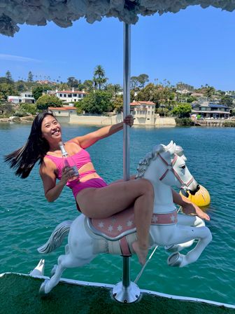 Smiling woman in a pink swimsuit rides a white carousel horse mounted on a boat, holding a bottled drink above blue bay water with waterfront homes and palm trees on a sunny day.
