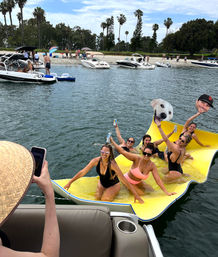 Group of women in swimsuits laughing and holding drinks on a bright yellow floating mat near anchored boats off a palm‑lined beach, a person in a straw hat photographing from a boat, with oversized dog and human face cutouts visible.
