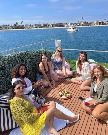 Six friends enjoying a sunny boat-deck picnic with wine and charcuterie, anchored in a calm beachfront harbor with yachts and coastal homes