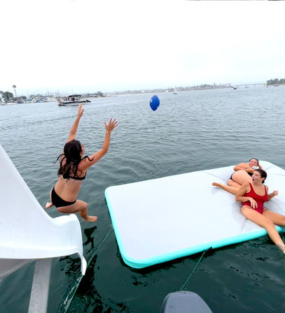 Three people in swimsuits playing on a white inflatable water mat beside a boat slide in a calm marina bay, one mid-air jumping toward a blue ball with boats and distant shoreline in the background.