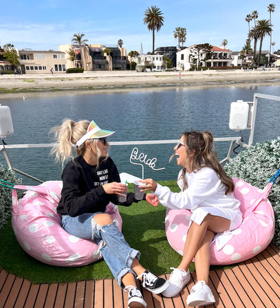 Two women laughing and toasting drinks on pink polka-dot bean bag chairs by a sunny coastal canal with palm trees and beachfront homes
