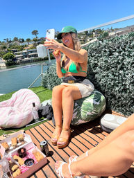 Person in a green bikini top and cap taking a selfie on a sunny waterfront deck, seated on a tropical-print beanbag with pastries and drinks nearby, bay water and hillside homes in the background and another friend’s legs in the foreground.